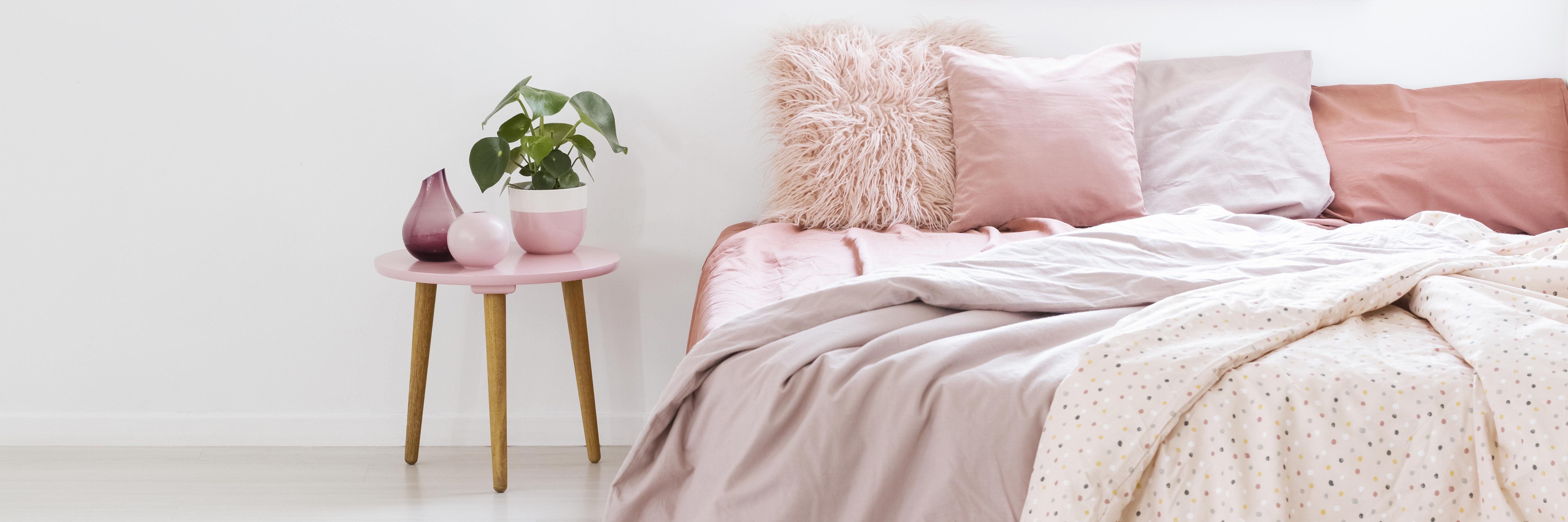 small table with a plant standing next to a bed with pink bedding in bedroom interior with white walls